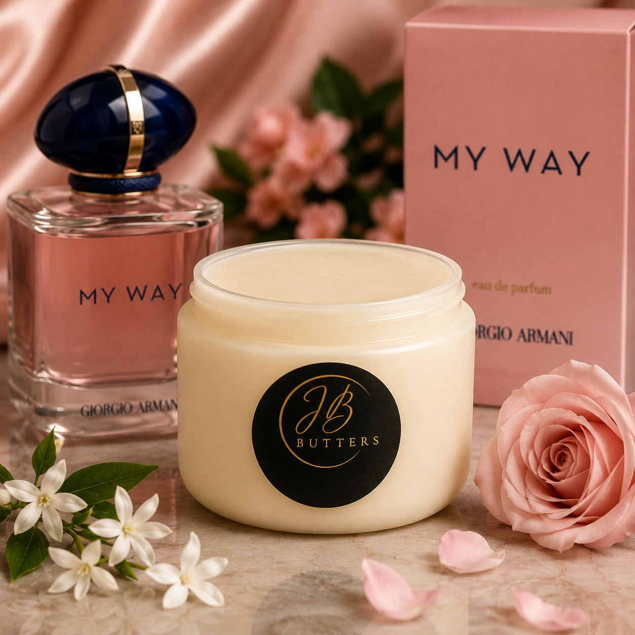 A jar of JB Butters body butter in a cream-colored container with a black circular label, styled alongside a Giorgio Armani MY WAY perfume bottle, a pink product box, white flowers, and a pink rose on a marble surface.
